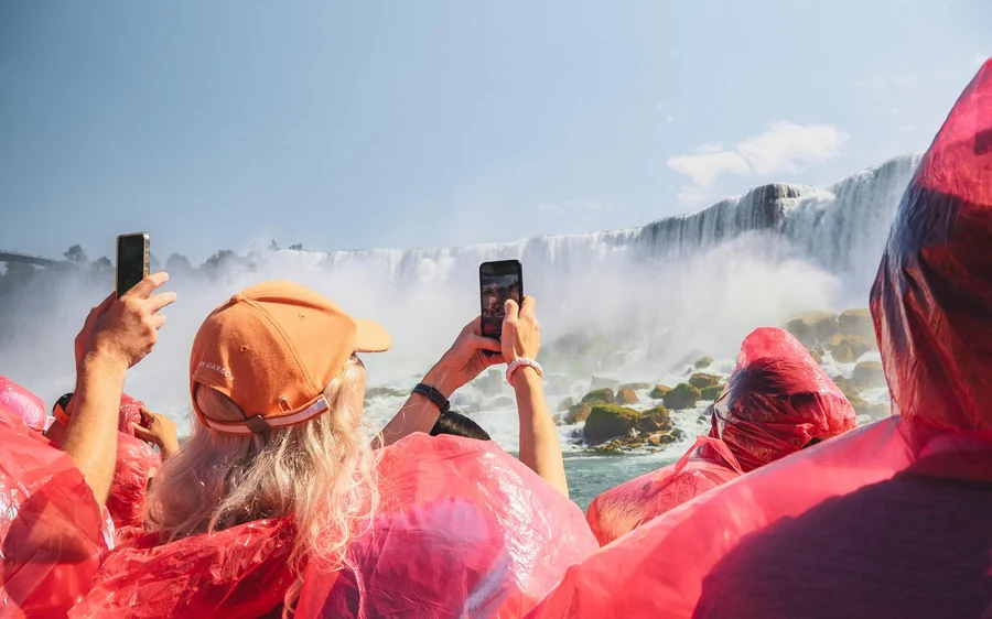 Passengers in red ponchos on a crowded open deck holding up phones in the mist