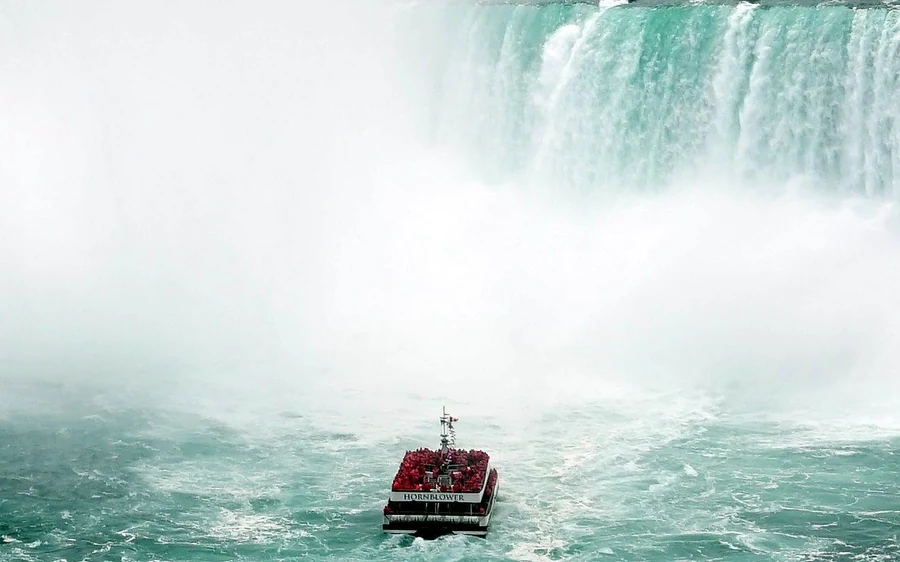 A small red boat far below a broad white wall of falling water and spray