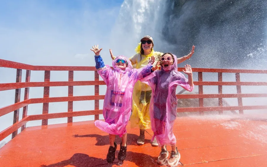 Three visitors in bright ponchos with their arms raised on a red deck below falling water