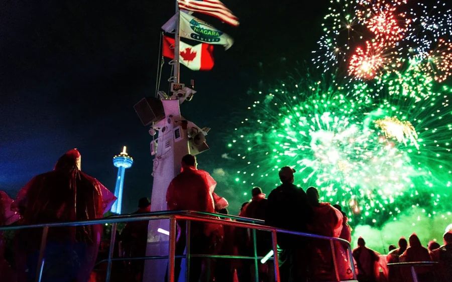 Green fireworks bursting over an illuminated boat deck at night with a Canadian flag on the mast