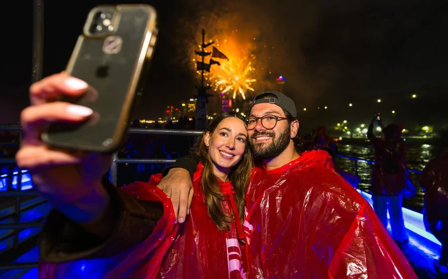 A couple in red ponchos taking a selfie at night with fireworks over the water behind them