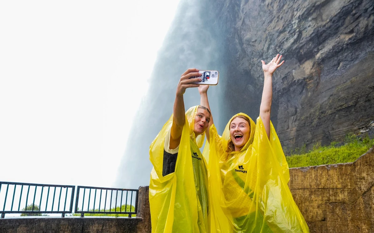 Two visitors in yellow ponchos taking a selfie at a railing with falling water behind them